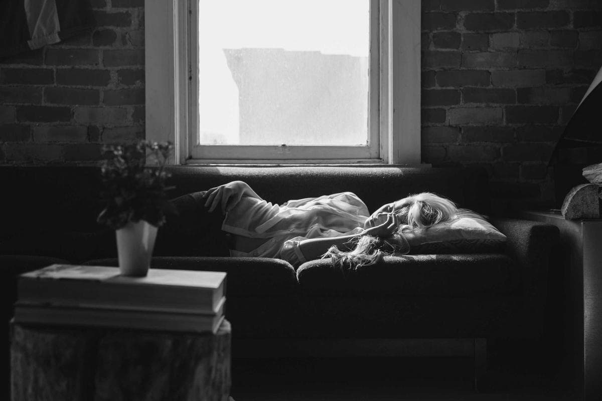 A person lying on a bed beside a sunlit window with a small potted plant on the sill, photographed in soft black and white.