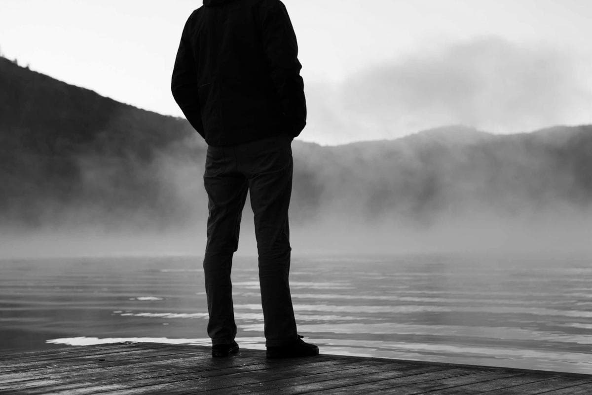 A lone figure standing at the edge of still water, facing into a thick bank of fog.