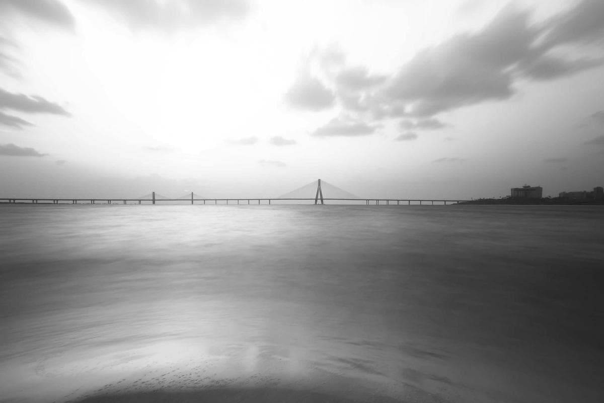 A long suspension bridge stretching across open water beneath a hazy sky, photographed in black and white.