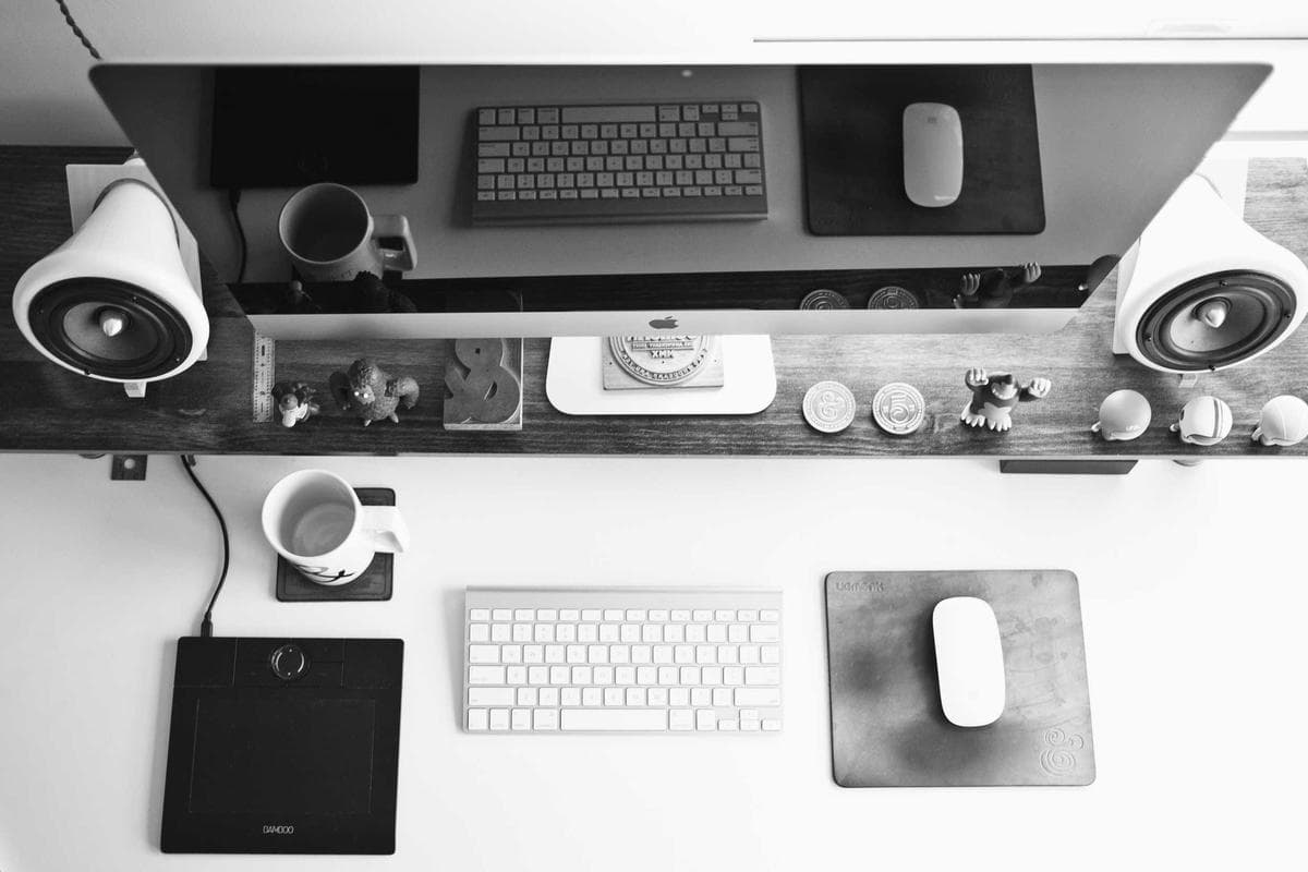 A desk workspace viewed from above, showing a widescreen monitor, speakers, keyboard, and mouse arranged neatly in grayscale.