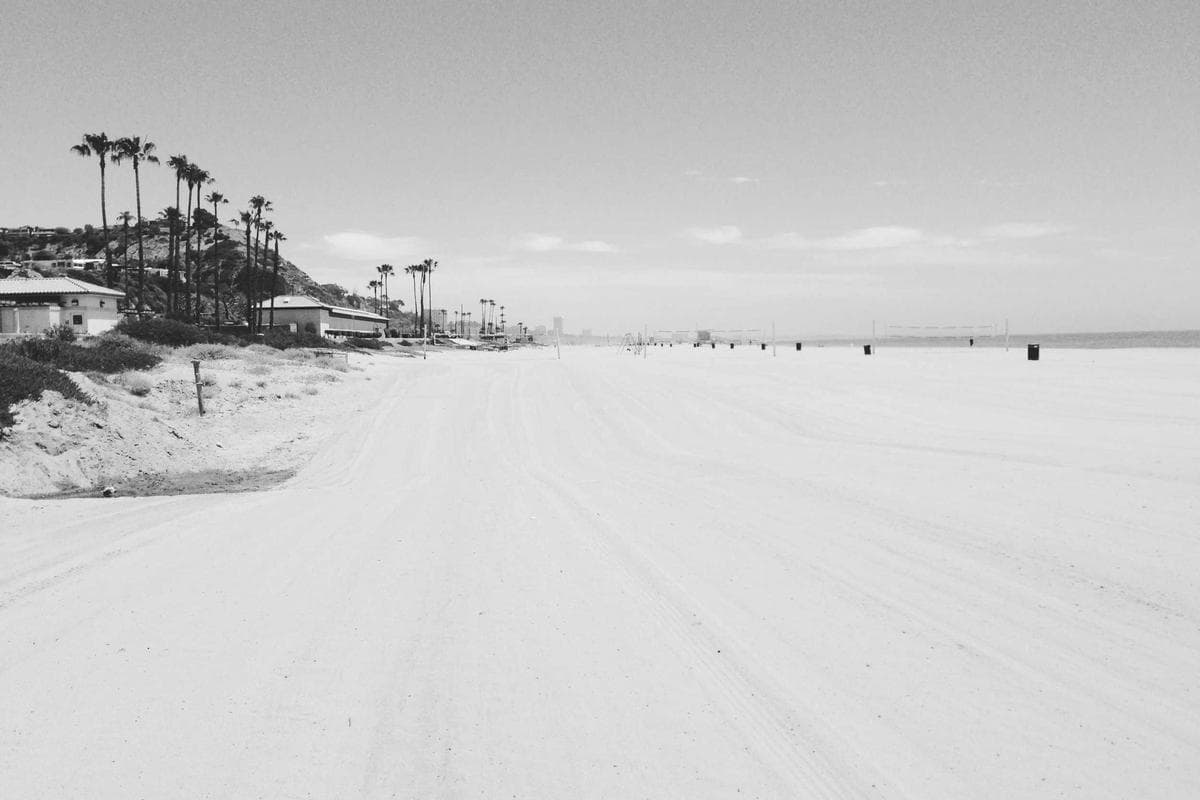 A vast, empty stretch of pale sand meeting a hazy horizon, with a few distant structures barely visible.